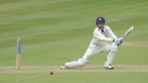 Amy Jones holding a cricket bat, wearing cricket whites, pads, and a helmet. There are also wickets and a ball on the grass. Picture credit: Matt Bright